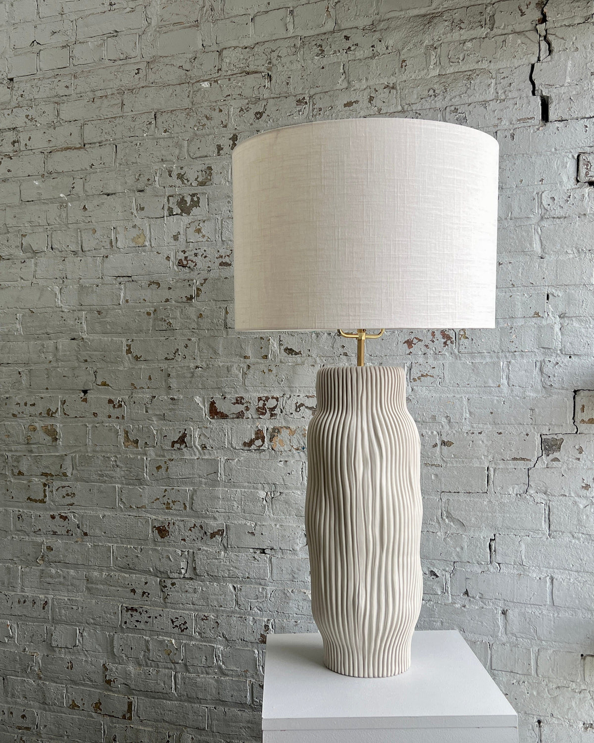 Della table lamp with cream porcelain base and off-white linen shade, displayed against a textured brick wall.