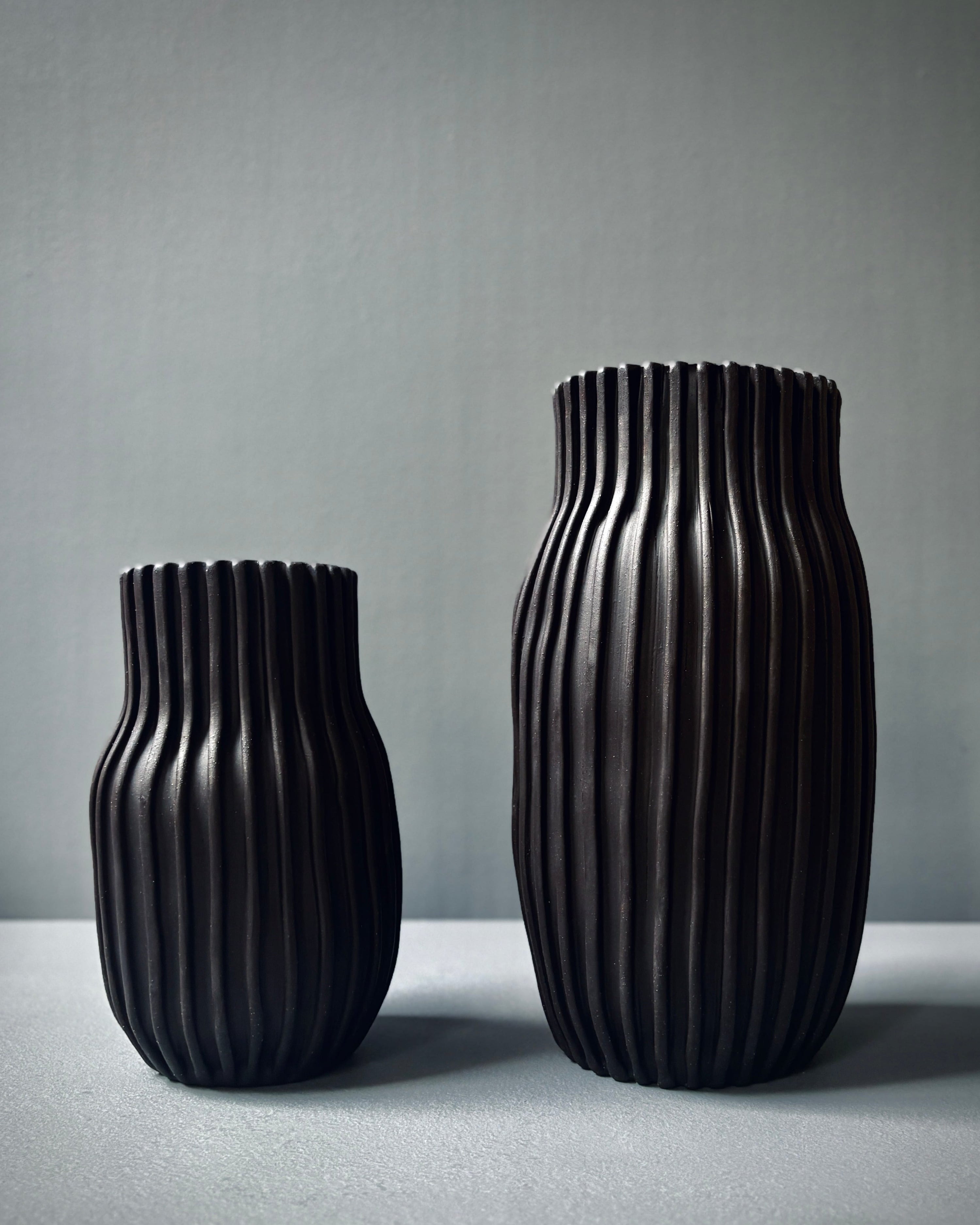 Two ribbed black ceramic vases of different heights standing on a light gray surface against a gray background.