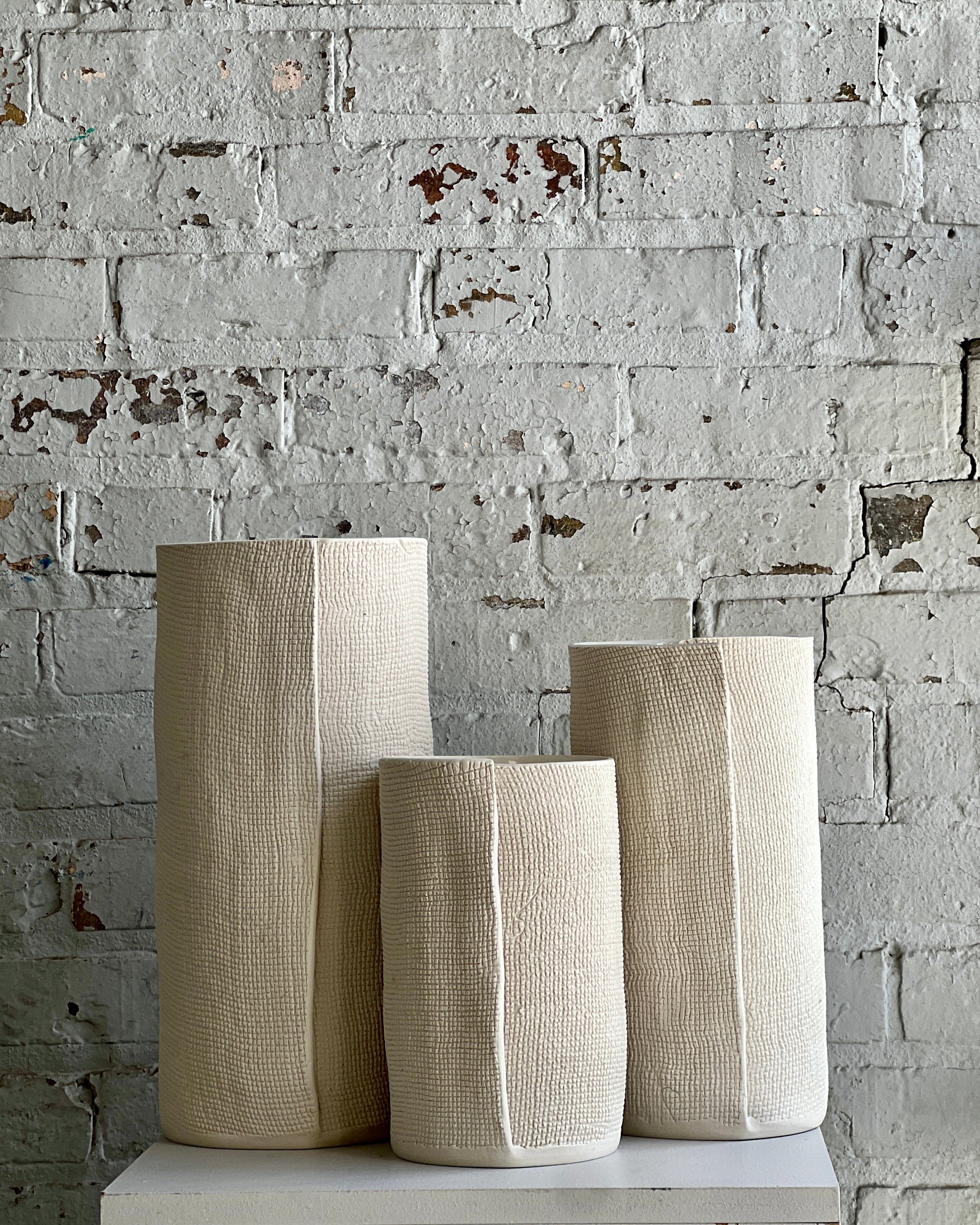 Three neutral-toned vases against a white brick wall.