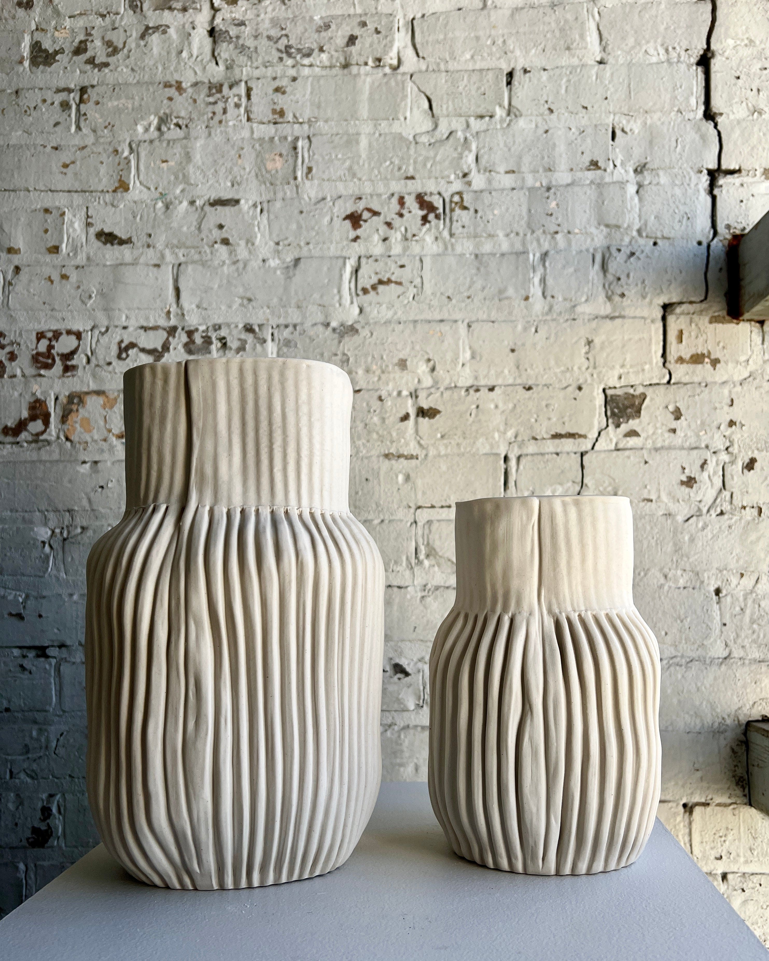 Two textured white vases on a table in front of a brick wall.