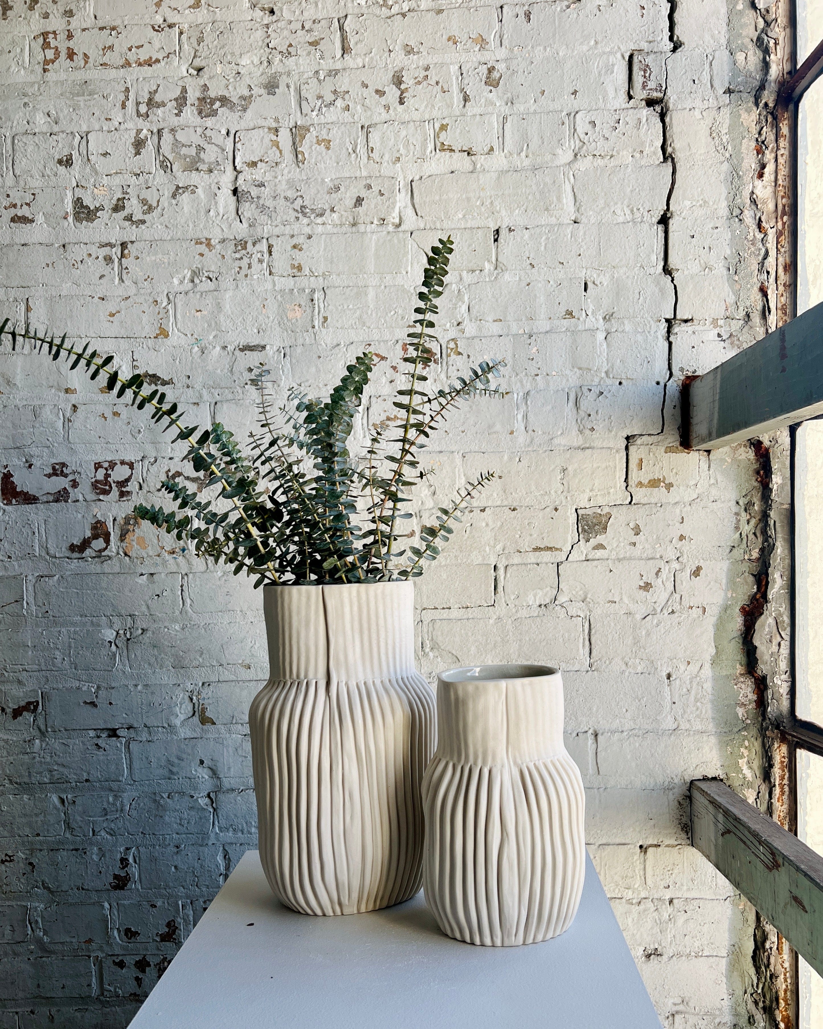 Two white vases with plants against a white brick wall.